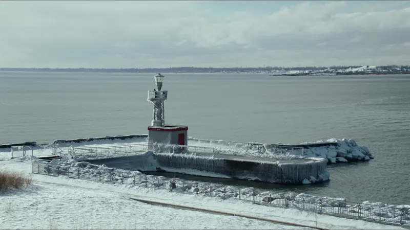 An iced over harbor pier with a small red doored light tower at its end, railings glazed in ice, a grey lake behind