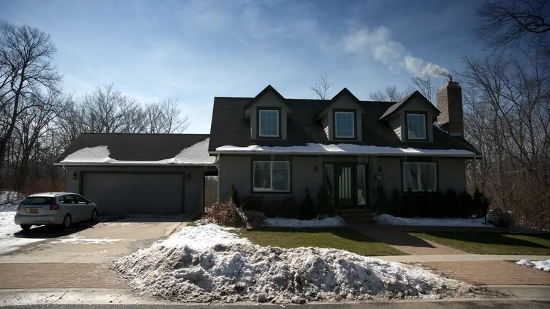 A grey Cape Cod house after a snowfall under a blue sky, smoke rising from the chimney, a car in the plowed drive