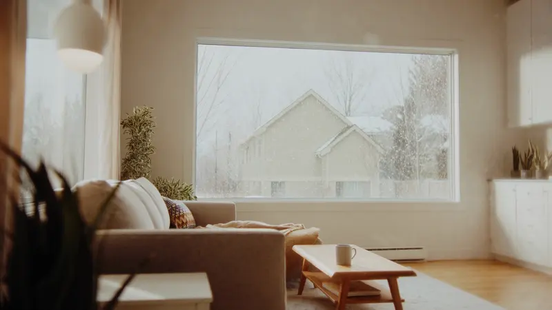 A bright living room with a big window, snow falling past the glass and a neighbouring house beyond, a mug on the coffee table