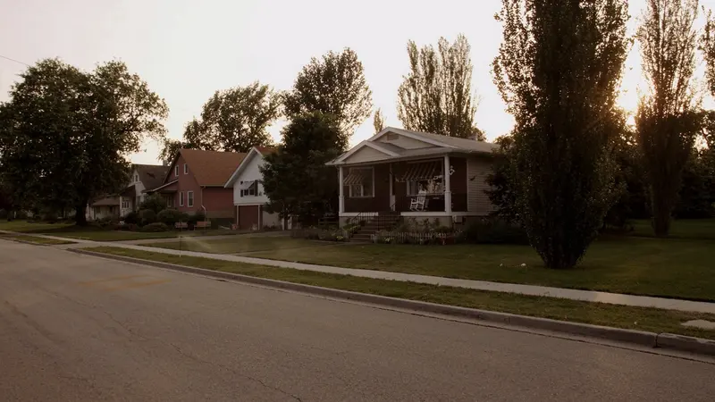 A quiet street of brick bungalows on a hazy July evening, a white rocking chair on a porch under striped awnings