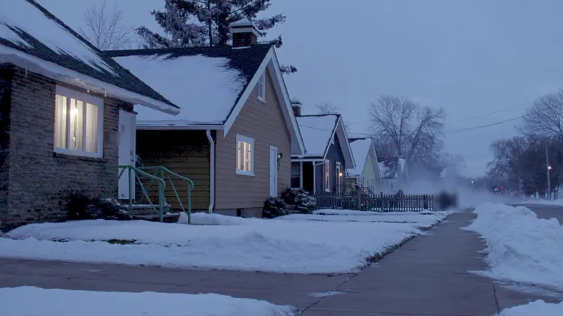 A snowy street of small houses at blue dusk, windows lit, a shoveled sidewalk running past them